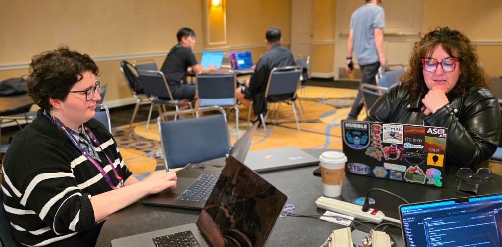 Two people collaborating on laptops at a round table during a PyCon US coding sprint. One person wears a PyCon US 2024 lanyard. The laptop on the right is covered in stickers for NumPy, NumFocus, Hubble, and other scientific open source projects.
